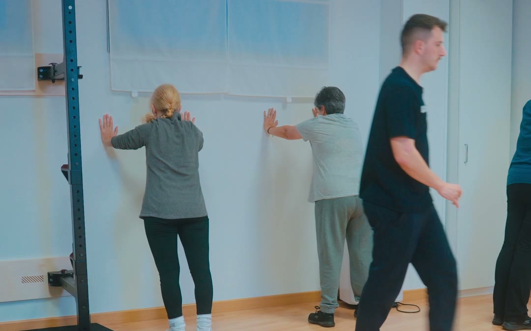 Dos personas trabajando apoyadas en la pared mientras un fisioterapeuta recorre la sala