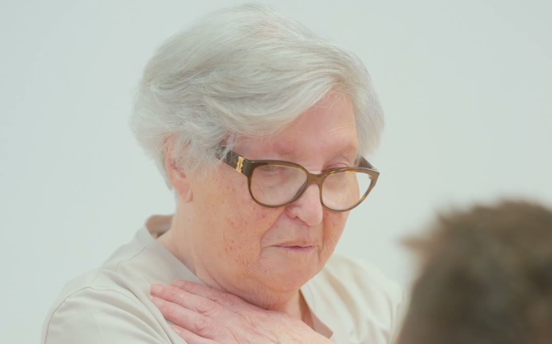 Primer plano de una mujer mayor con la mano apoyada sobre el pecho, con un fisioterapeuta a su lado