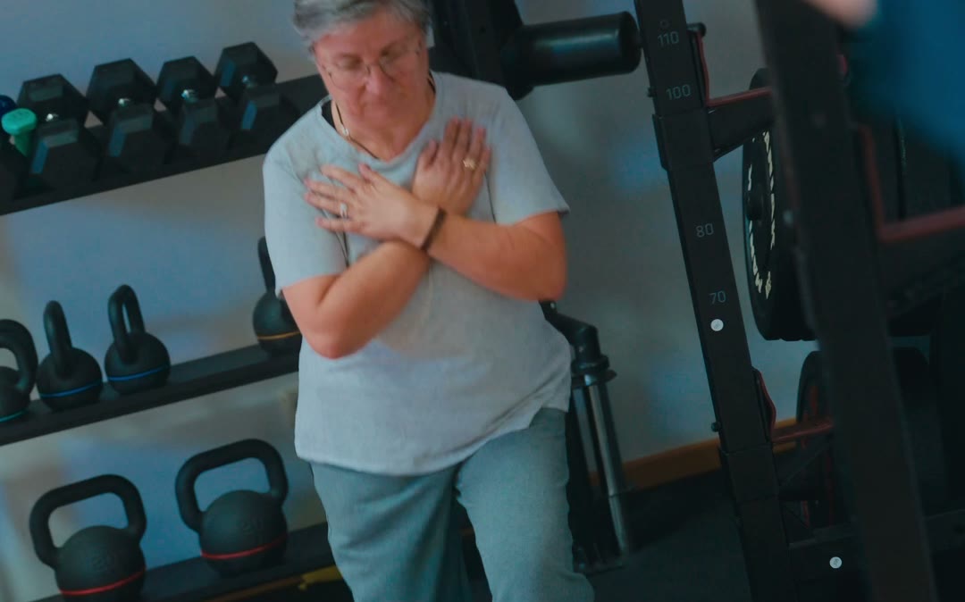 Mujer con los brazos cruzados sobre el pecho levantándose, con las pesas de la sala detrás