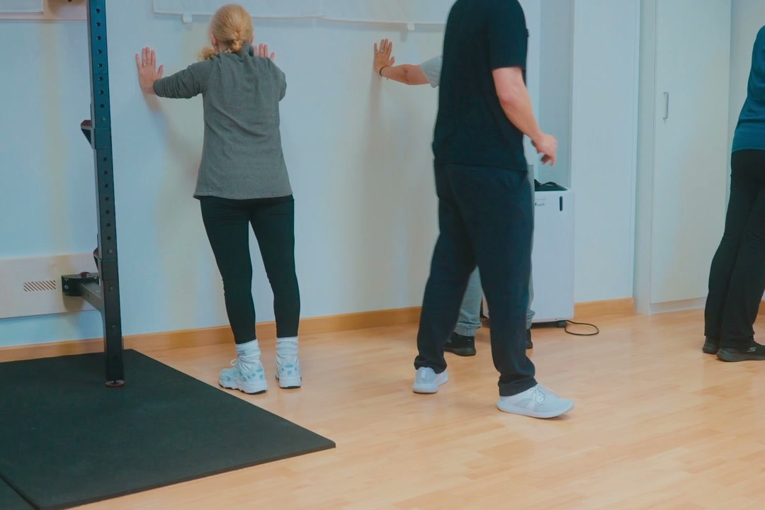 Grupo haciendo un ejercicio de brazos frente a la pared, guiado por un fisioterapeuta