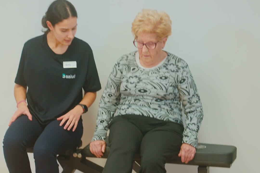 Fisioterapeuta sentada en el banco junto a una mujer mayor, las dos de frente a la sala
